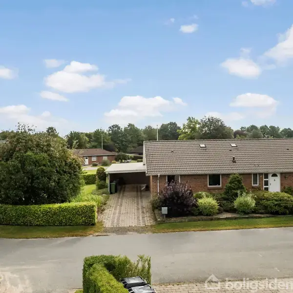 En enfamilieshus med carport står ved en vej i et grønt villakvarter. Det har en murstensfacade, velplejede buske foran, og omgivelserne er rolige med træer.
