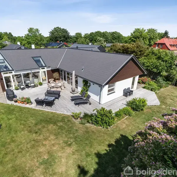 Hus med mørkt tag og store vinduer står på en græsplæne med terrassemøbler. Det er omgivet af en have med buske og træer i et villakvarter.
