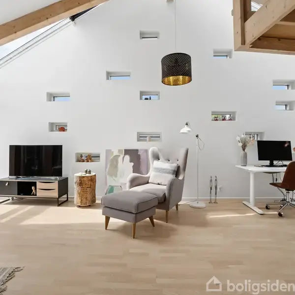 Stue med tv-bord, lænestol og skrivebord i lyse omgivelser. Skrålofter med ovenlysvinduer, hvidmalede vægge og lyse trægulve. Grøn udsigt gennem glasdør til venstre.