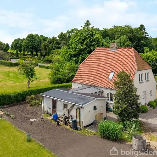 Et hvidt hus med rødt tag står i en frodig have omgivet af træer og buske. I forgrunden ses en lille bygning med et fladt tag.