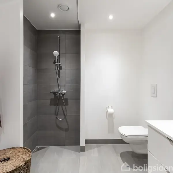 Et moderne badeværelse med en grå flisebrusekabine til venstre. Hvide vægge, et væghængt toilet, håndklæder på kroge, samt en kommode med spejl og diverse småting.
