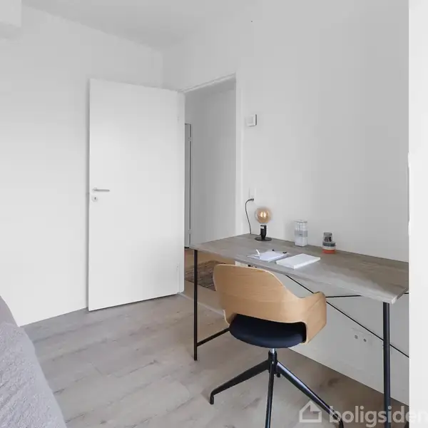 Et skrivebord med kontorstol og lampe står ved en væg. Papirer og en bog ligger på bordet. Til venstre ses en seng i et lyst rum.