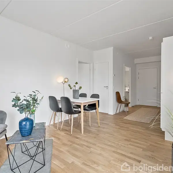 Gråt sofaarrangement står i en stue med trægulv; spisebord med fire stole i baggrunden. Enkel, moderne indretning med indendørsplanter og væglamper. Dør i baggrunden leder til gang.