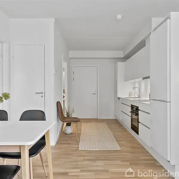 Et moderne køkken med hvide skabe står langs væggen. Et spisebord med sorte stole står i forgrunden. Trægulv og en gulvløber fører til en hvid dør.