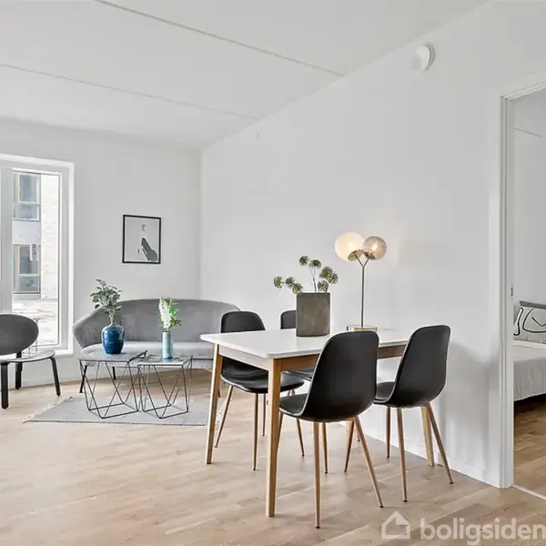 Stue med lyse træmøbler og sorte stole omkring et spisebord, grå sofa og runde sofaborde, placeret nær et stort vindue. En dør fører til et soveværelse.