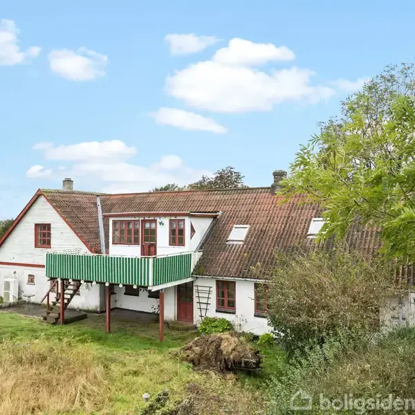 Stuehuset med rødbrun tag, grøn altan og hvid facade står stille i landligt område med græs og træer omkring.