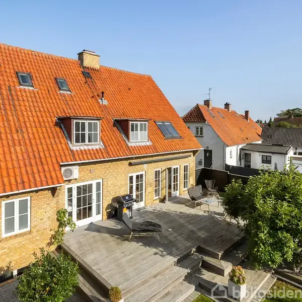 Et gult murstenshus med rødt tegltag har en solrig terrasse. Huset har flere små vinduer på taget, der vækker en hyggelig atmosfære. Terrassekanten er omgivet af grønt.