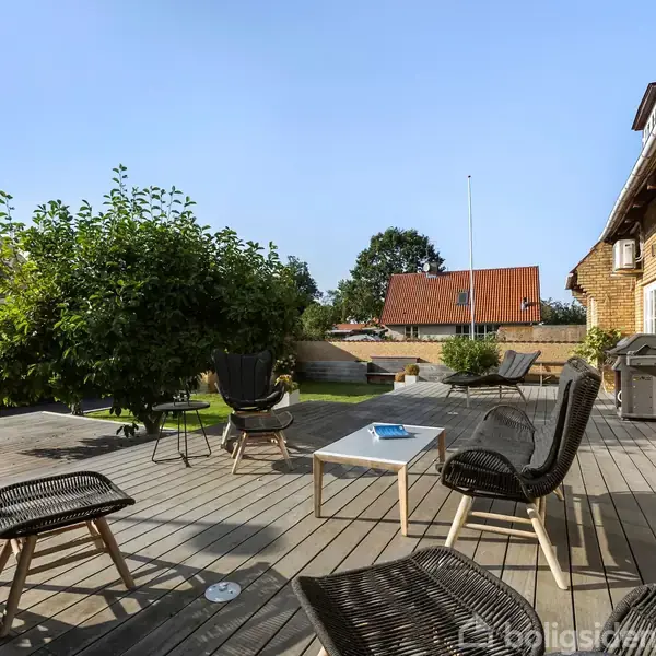 Terrassemøbler står på en træterrasse foran et hus med murstensvægge. I baggrunden ses en have med et træ og flere huse med røde tage. Skyfri himmel.