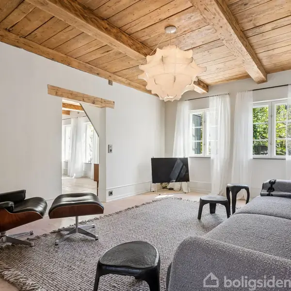 Stue med en grå sofa og en sort lænestol, placeret ved et stort vindue. Rummet har et træloft og en neutral farvepalet med en stor, lys lampe i loftet.