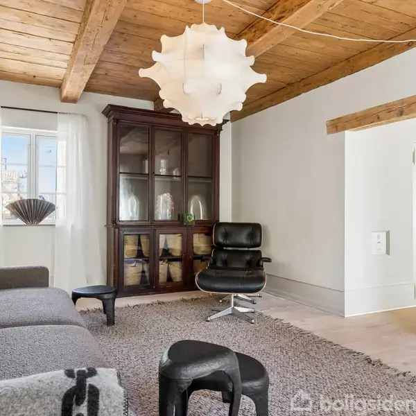 En stue med en grå sofa står mod venstre, ved siden af et sort lænestol på et tæppe. En mørk vitrineskab står ved vinduet. Loftet har træbjælker og en skulpturel lampe.