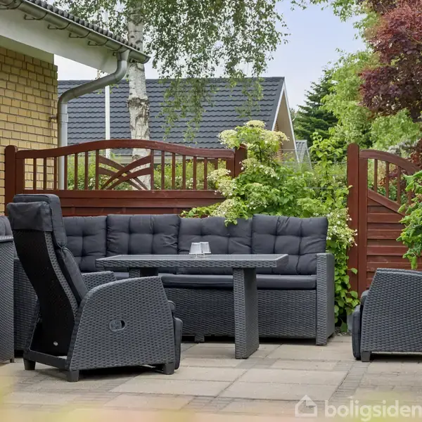 Mørkegråt udendørs loungesæt med sofa og stol omkring et bord. Placeret på terrasse ved gul teglstensmur og træhegn, omgivet af buske og træer.