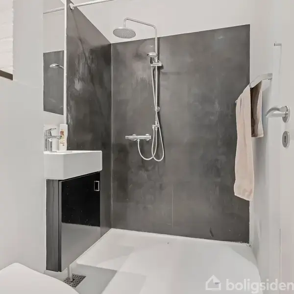 Badeværelse med sort vægpanel, bruser monteret på væggen, og hvid håndvask med dispenser. En beige håndklæde hænger til højre. Moderne design med enkelt indretning.