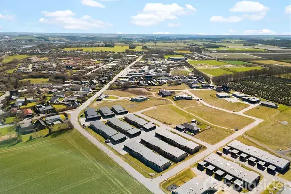 Luftfoto viser et boligområde med rækkehuse omgivet af åbne marker og grønne områder. Byens infrastruktur med veje og mindre bygninger kan ses i baggrunden under en blå himmel.