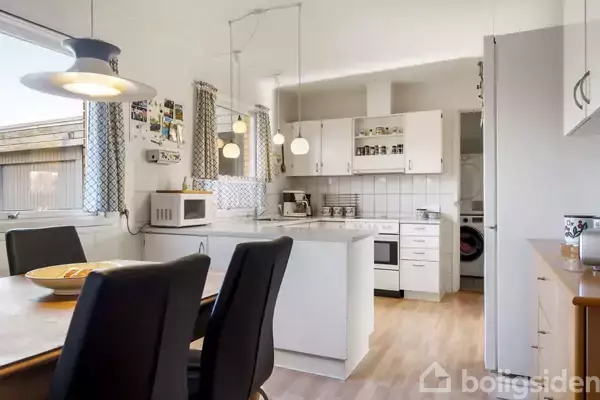 Et køkken med hvide skabe og apparater, inklusive en mikroovn og en ovn, omgivet af lyse træmøbler. Der hænger lamper over spisebordet med sorte stole.