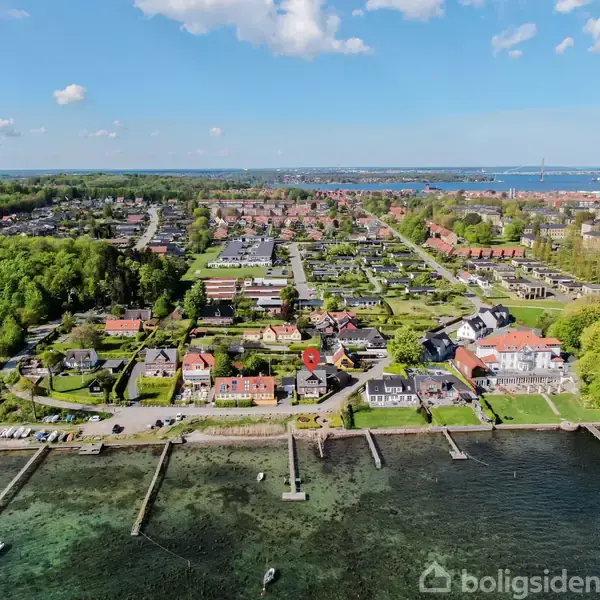 Luftfoto viser en række huse langs en kystlinje med moler ud i vandet. Grønt landskab omkranser bebyggelsen, og en by strækker sig i baggrunden under en blå himmel med få skyer.