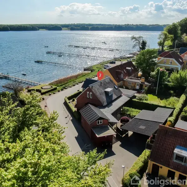 Rødt hus med sort tag ved vejen i en landsby. Omgivet af grønne haver og skov. Nær en bredde med udsigt til sø og skyer i baggrunden.