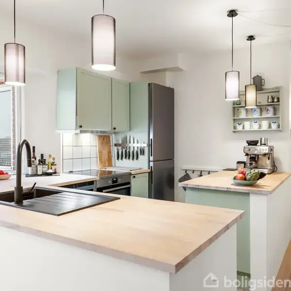 Køkken med pastelfarvede skabe og moderne apparater; en vask og kogeplade er på en lys træbordplade. Omkring hænger lamper, mens hylder rummer køkkenting. Spisebord ses i baggrunden.