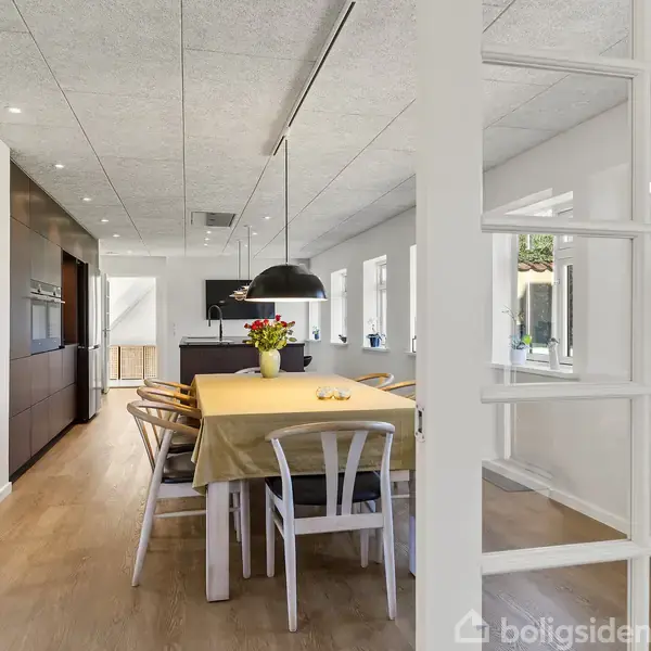 Spisebord dækket med en dug og blomster, omgivet af stole i et lyst køkken med trægulv og store vinduer; hængelamper ovenover, glasdør til højre, grøn have udenfor.