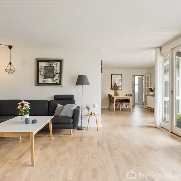 En stue med en mørkegrå hjørnesofa ved et sofabord, udsmykket med blomster. En gulvlampe står ved siden af, og der er store vinduer med udsigt til en terrasse.