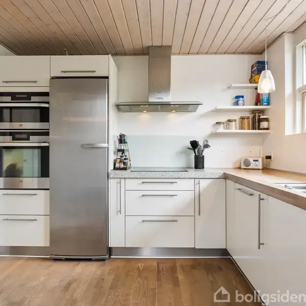 Køkken med hvide skabe, stålkøleskab og indbyggede ovne på venstre side. Trægulv og træloft. Køkkenredskaber står ved vask på højre bordplade. Store vinduer giver naturligt lys.
