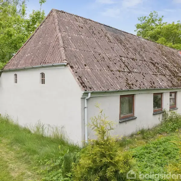Et hvidt landhus med et gammelt, brunt tag står omgivet af frodig vegetation og græs. Vinduerne er små og traditionelle. Det naturlige landskab dominerer omgivelserne.