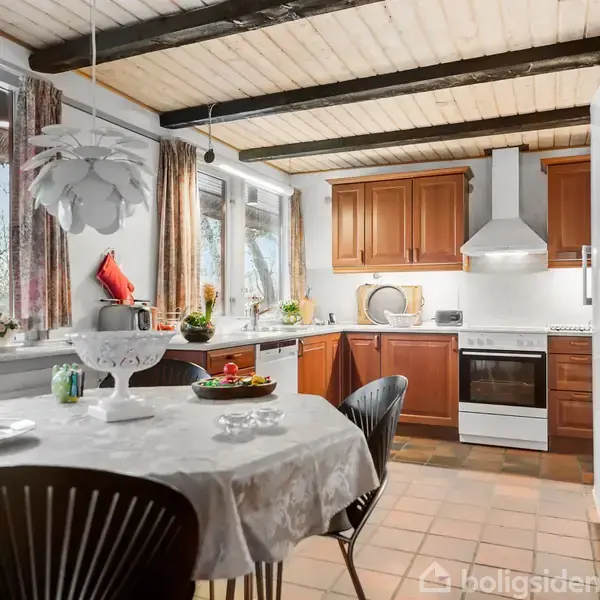 Et køkken med brune skabe og en hvid komfur-emhætte. Et bord med en hvid dug og stole står midt i rummet. Store vinduer giver udsigt til træer udenfor.