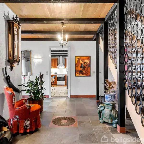 En entré med en rød stol står foran en spejlvæg, omgivet af planter. Til højre en trappe med dekorative sorte gelændere. I baggrunden ses åben dør til køkken.