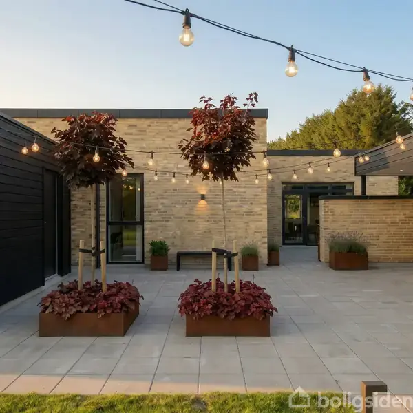 En moderne gårdhave med to dekorative plantekasser med træer. Lyskæder er spændt over gårdhaven, hvor der ses et hus i baggrunden med brune murstensvægge og sorte detaljer.