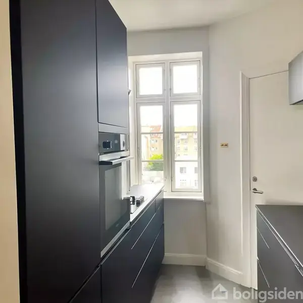 Moderne køkken med sorte skabe og integreret ovn, set fra siden, hvor dets blanke overflader reflekterer lys fra et stort vindue, der giver udsigt til bybebyggelse udenfor.