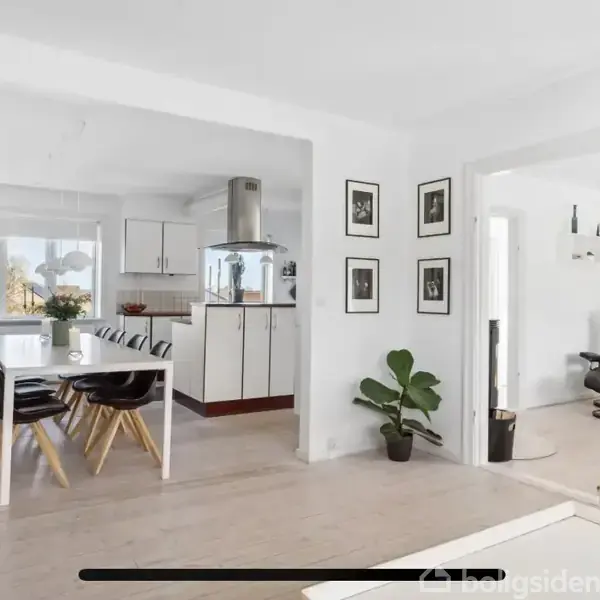 Spisebord med sorte stole står i et lyst køkken. I baggrunden er der hvide skabe og en stålventilator. Stue med lænestol ligger ved siden af, adskilt af en væg med billeder.