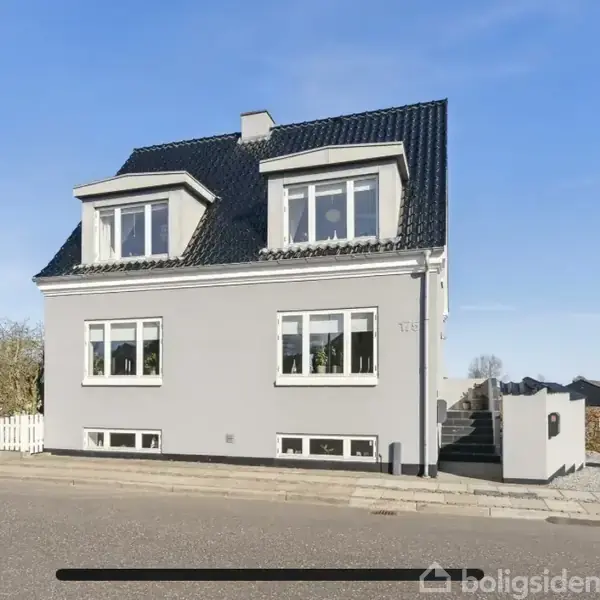 Grå murstenshus står stille ved vejen med sort tag og hvidrammede vinduer, omgivet af et hegn og træer i et beboelseskvarter.