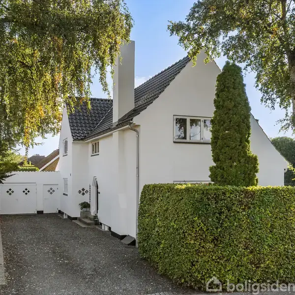 Hvidt hus med mørkt tag og skorsten, omgivet af frodige træer og hække. Indkørsel fører til huset med en havelåge i baggrunden. Solrigt vejr skaber en lys atmosfære.