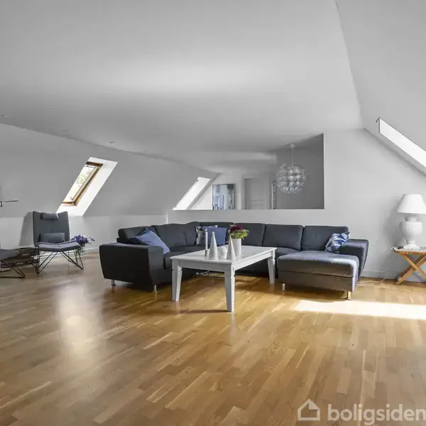 Grå sofa med sofabord står i en lys, rummelig stue med skråvægge og trægulv. Flere vinduer lader sollys strømme ind. To stole og en lampe ses i baggrunden.