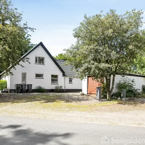 Et hvidt hus med tag af mørke tegl står roligt omgivet af grønne træer i en landlig setting. En rød garageport drejer ud mod en stille vej.