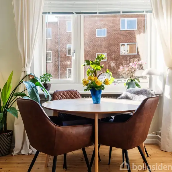 Et rundt bord med brune stole står foran et vindue. En vase med gule blomster pryder bordet. Omkring er grønne planter og gardiner, der giver et hyggeligt hjemmemiljø.