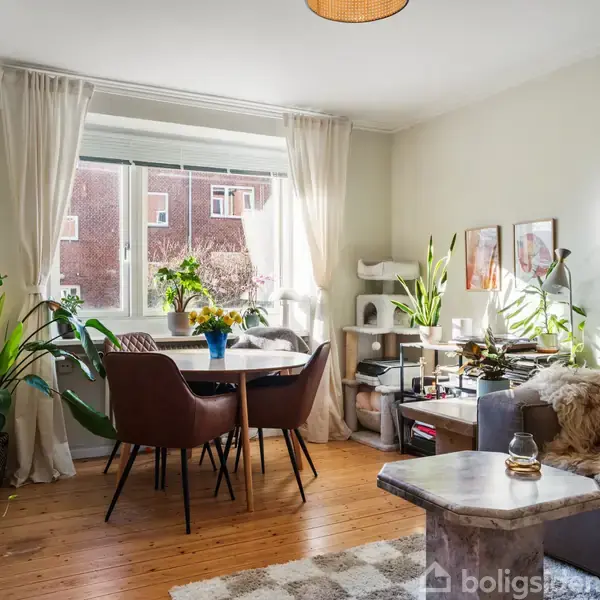 Stue med sofa, sofabord og spisebord, omgivet af grønne planter. Store vinduer lukker lys ind. Væggene har kunstværker, og der er en hyggelig stemning.