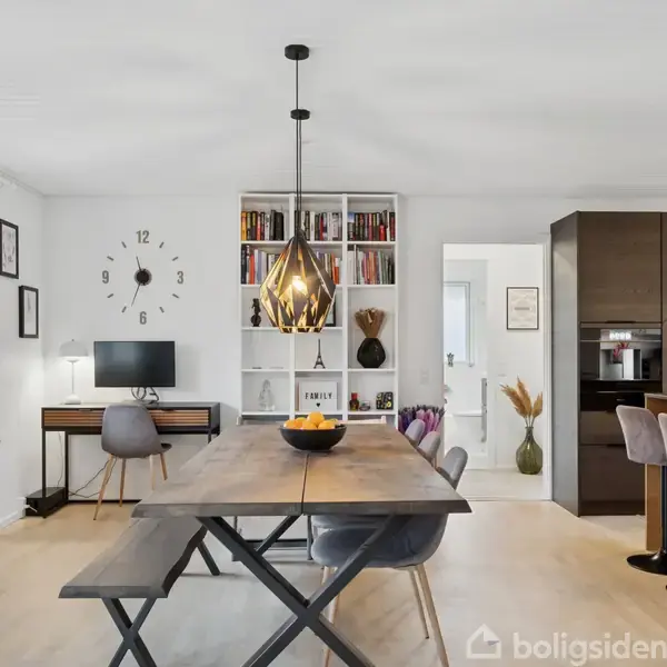 Et træbord med stole står i en moderne stue. Over bordet hænger en dekorativ lampe. Bagved ses en reol med bøger, et skrivebord med computer og en vægklokke.