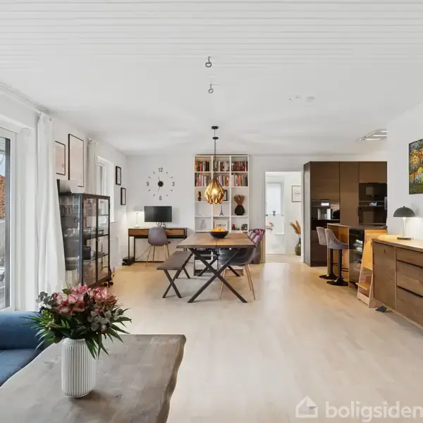 En stue med et blåt sofaområde har et sofabord med planter. Spisebordet står midt i rummet under en lampe, og køkkenet ses i baggrunden. Store vinduer giver udsigt til terrassen.