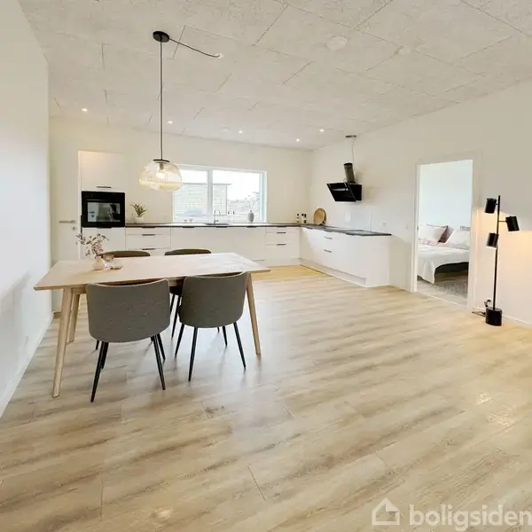 Køkken med spisebord, to stole under en loftslampe, i moderne hvidt rum med trægulv. I baggrunden ses køkkenelementer med ovn, vask og vindue, samt en åben dør til et tilstødende værelse.