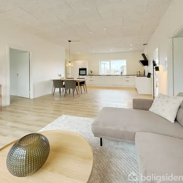 En lys stue med en beige sofa og et rundt sofabord med en dekorativ vase. I baggrunden ses et åbent køkken med spisebord og fire stole.