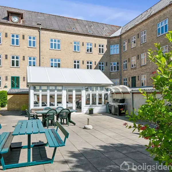 En gårdhave med bord og bænke i træ står stille på flisebelagt terrasse foran et drivhus, omgivet af en murstensbygning med flere vinduer. Grønt løv danner en naturlig ramme.