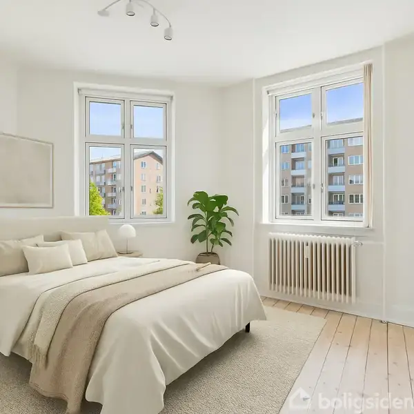 Soveværelse med dobbeltseng, sideborde og lamper, står i et lyst rum med trægulve. Planter og kunst pryder væggene ved siden af store vinduer med udsigt til et lejlighedskompleks.