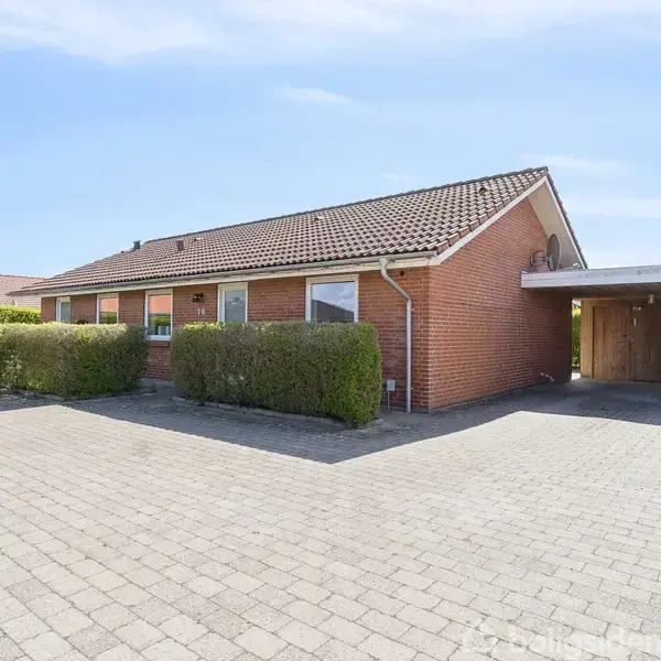 Et murstenshus med rødt tag står på en brolagt gårdsplads. Til højre er en carport, delvist dækket af buske. Himlen er klar og blå.