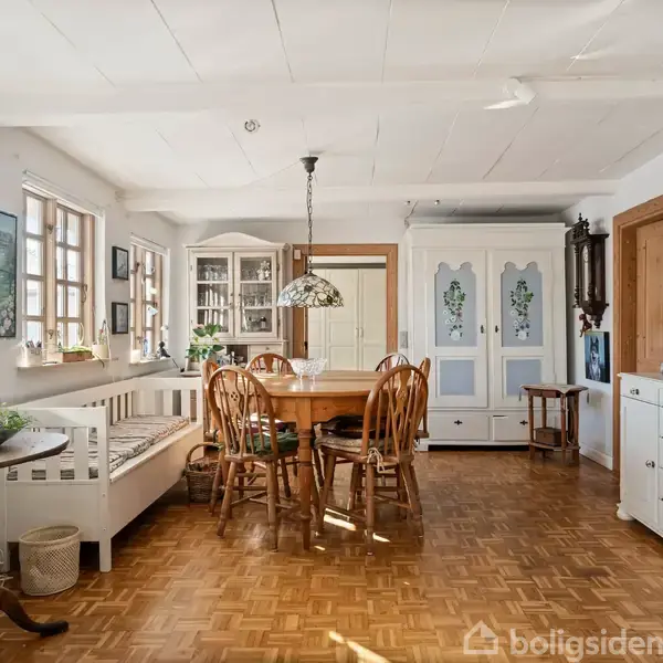 Spisebord med stole står i centrum af en lys spisestue. Vinduerne til venstre lader sollys stråle ind, og skabe samt en kommode pryder rummet. En bænk står ved vinduet.