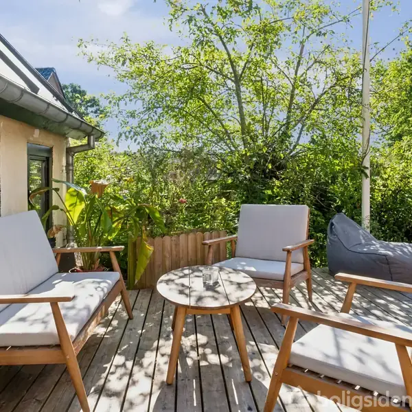 Tre udendørs stole og et rundt bord står på en træterrasse, omgivet af grønne buske og træer i solrig havekontekst. Huset ses delvist til venstre.