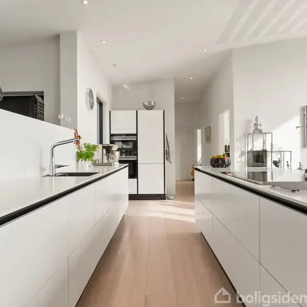 Et moderne køkken med hvide skabe og en vask i midten. Omgivelserne er lyse med trægulv, store skuffer og nogle dekorationer på bordpladen.