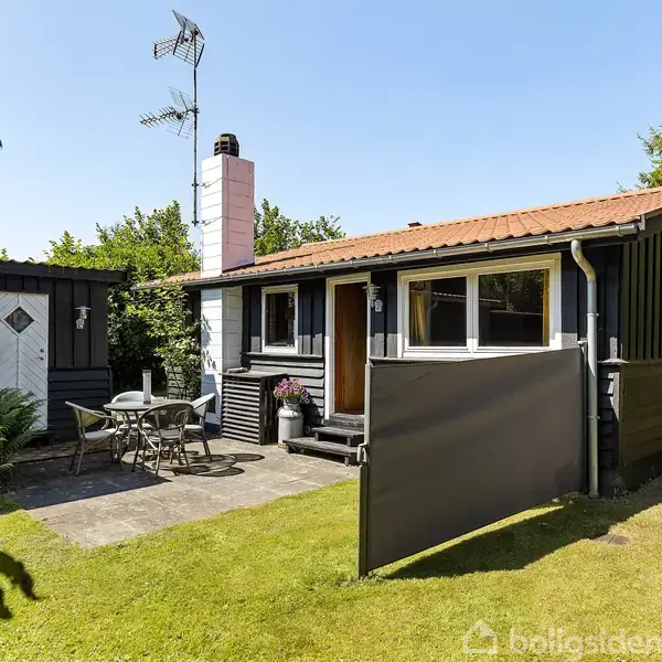 Sommerhus med sort træbeklædning og rødt tegltag. Terrasse foran huset har et cafésæt. Græsplæne omgiver huset, og en anden bygning ses til venstre. Solrigt vejr.