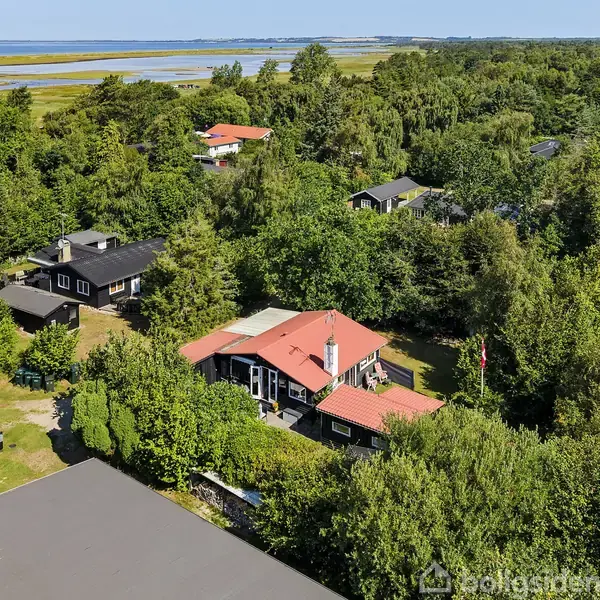 Hus med rødtag ligger i en frodig, skovklædt sommerhusområde nær kysten i baggrunden. Flere huse omgiver det, og der er grøn vegetation overalt.