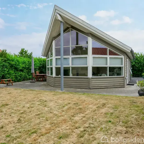 Hus i moderne design med store vinduer, der står på en græsplæne. Omkring huset er der terrasse, havemøbler og frodig hæk.