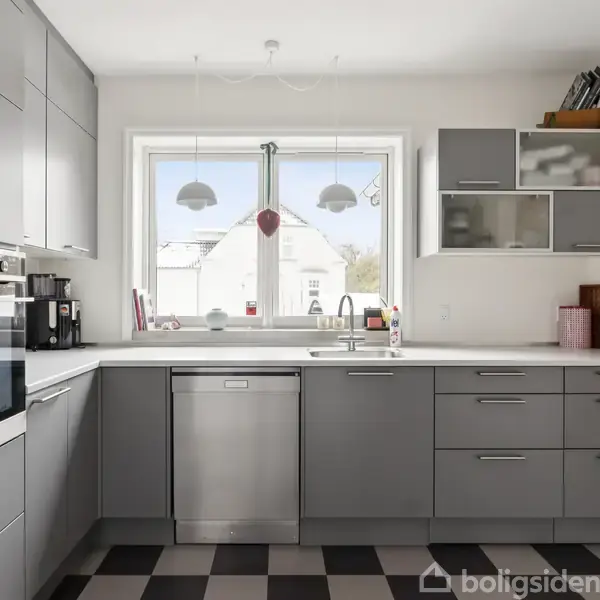Et moderne køkken med grå skabe og skuffer, en opvaskemaskine under et stort vindue, i en velbelyst rum med ternet sort-hvidt gulv. Elementer inkluderer kaffemaskine og skærebræt.